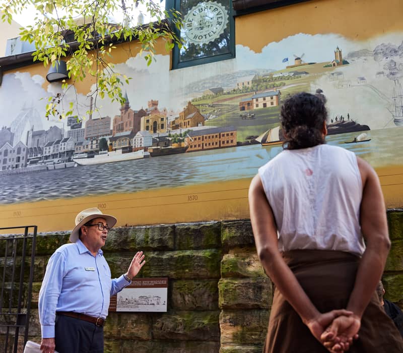 A tour guide talking to tourists in front of a historical mural at The Rocks in Sydney.