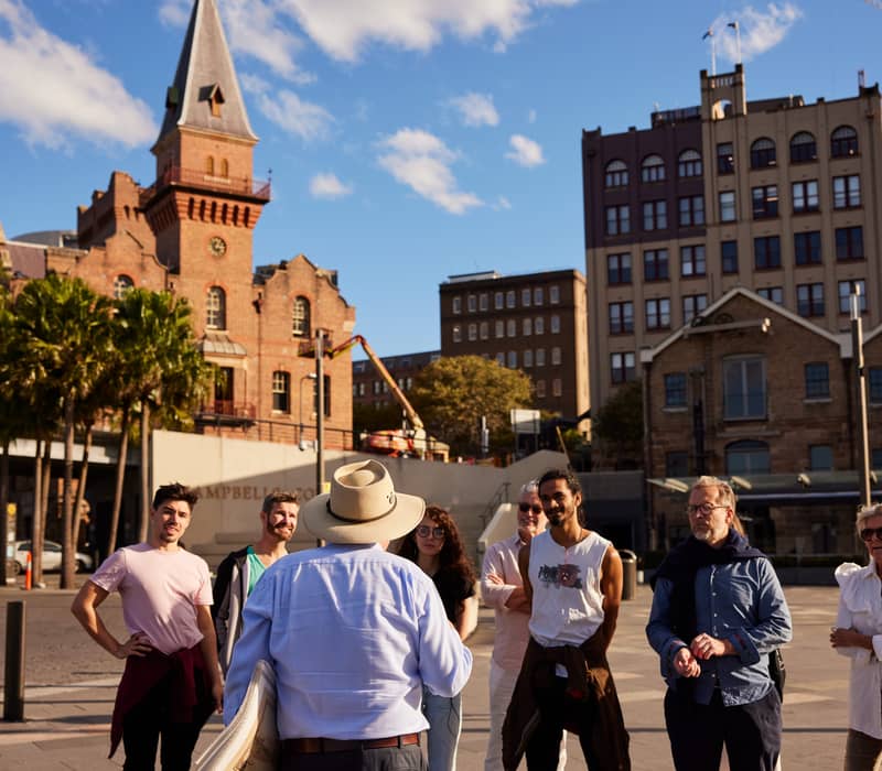 Tourists gathered in a public square at The Rocks in Sydney with historic brick buildings in the background.