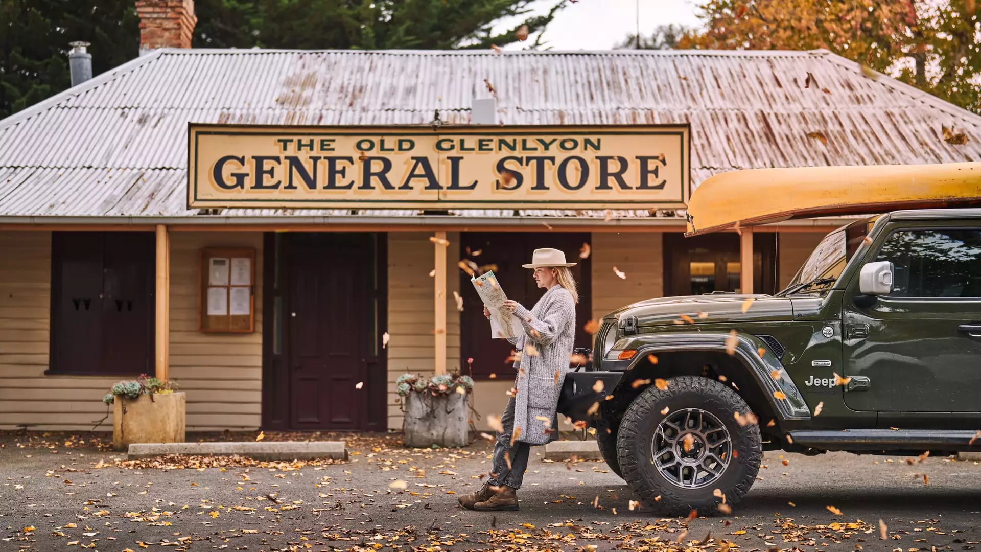 A woman in a hat leans against a 4WD with a canoe, reading a map outside a rustic general store.