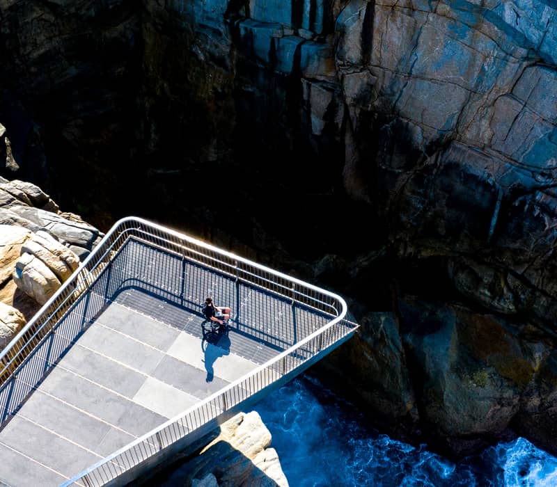 A high-angle shot of a person on the cantilevered grating platform at The Gap, overlooking crashing blue waves in Albany, WA.