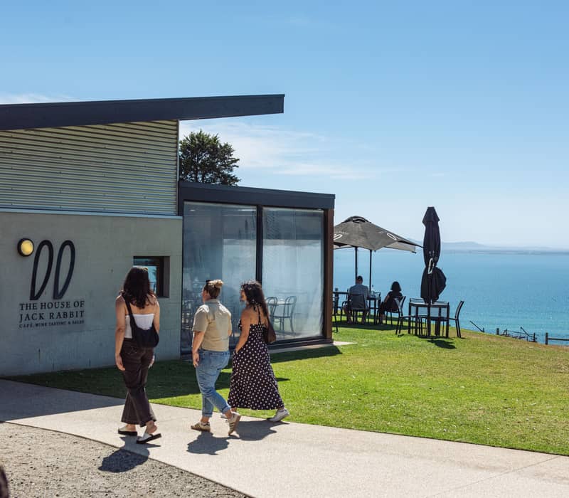 A modern industrial-style cellar door building with a large outdoor deck and people enjoying wine tasting overlooking the ocean.