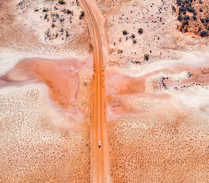 A top-down aerial photograph of a vibrant orange dirt road cutting a straight path through the pale, textured salt pans and arid shrubland of the Western Australian Goldfields.
