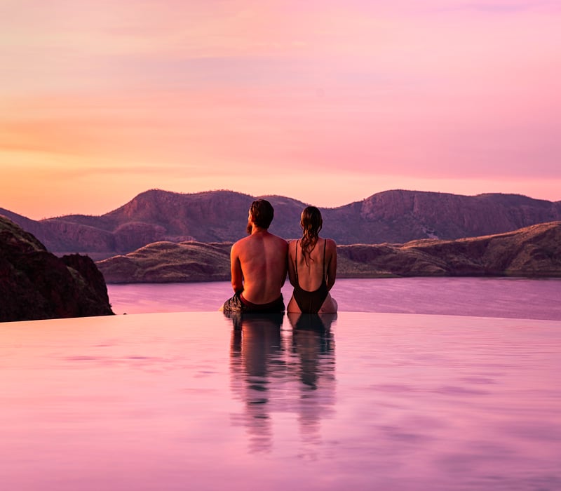 A couple relaxes at the edge of the famous infinity pool at Lake Argyle Resort, overlooking the vast, sunset-lit waters of the inland sea and the Carr Boyd Ranges.