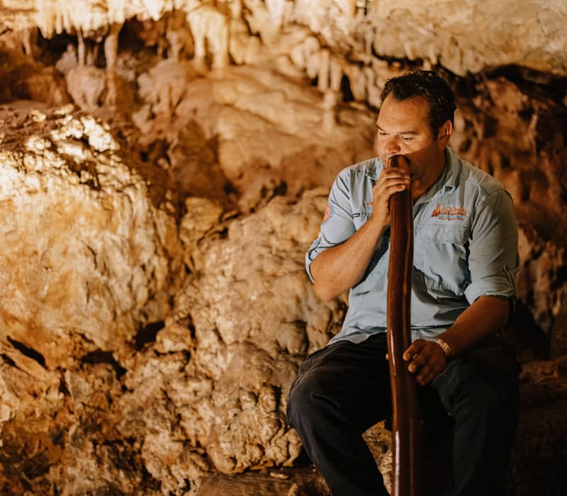 Close-up of an Aboriginal man playing a long wooden didgeridoo inside a textured limestone cave.