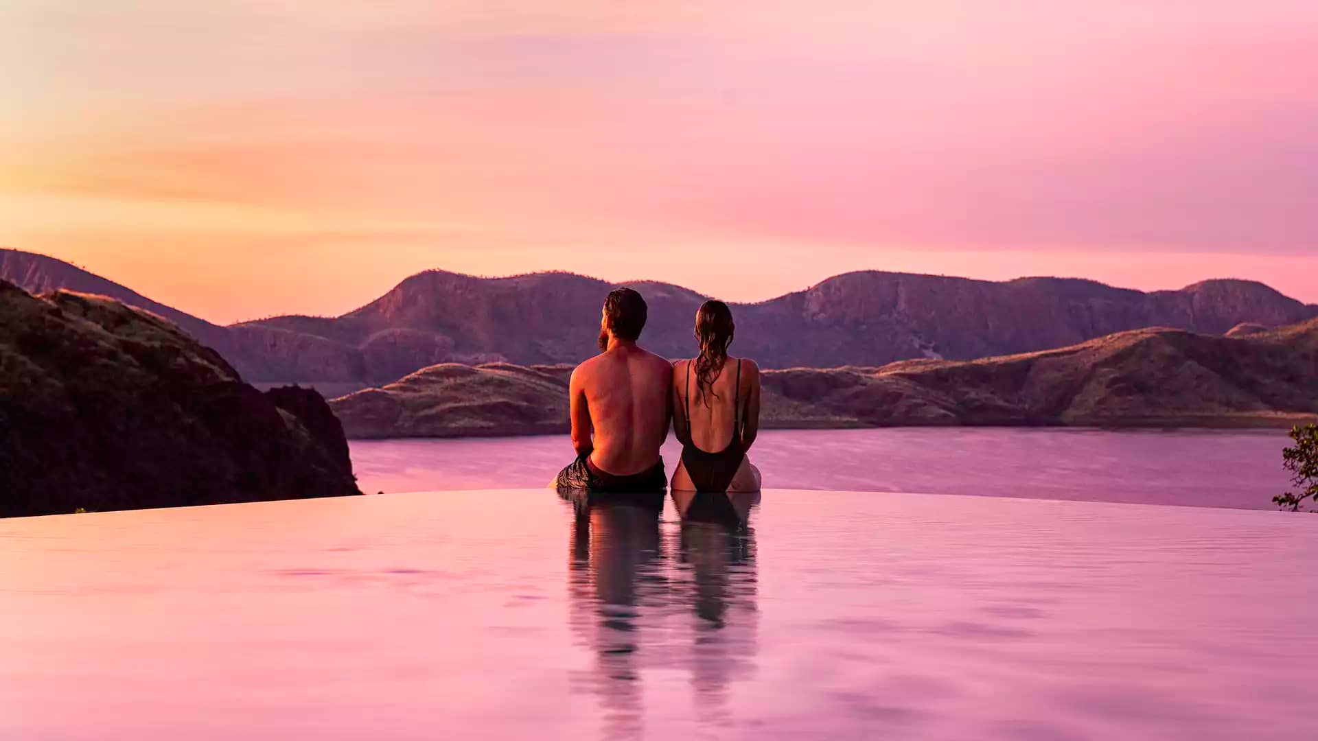 A couple relaxes at the edge of the famous infinity pool at Lake Argyle Resort, overlooking the vast, sunset-lit waters of the inland sea and the Carr Boyd Ranges.