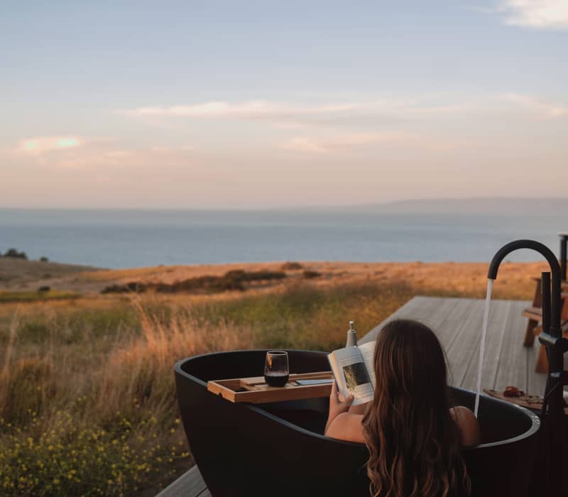 Woman reading a book in an outdoor bathtub at CABN X overlooking the coast.