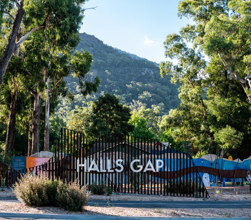 A vertical shot shows a stylish metal slat sign spelling "HALLS GAP" on a roadside in Victoria, backed by tall gum trees and a high, densely forested mountain under a clear blue sky.