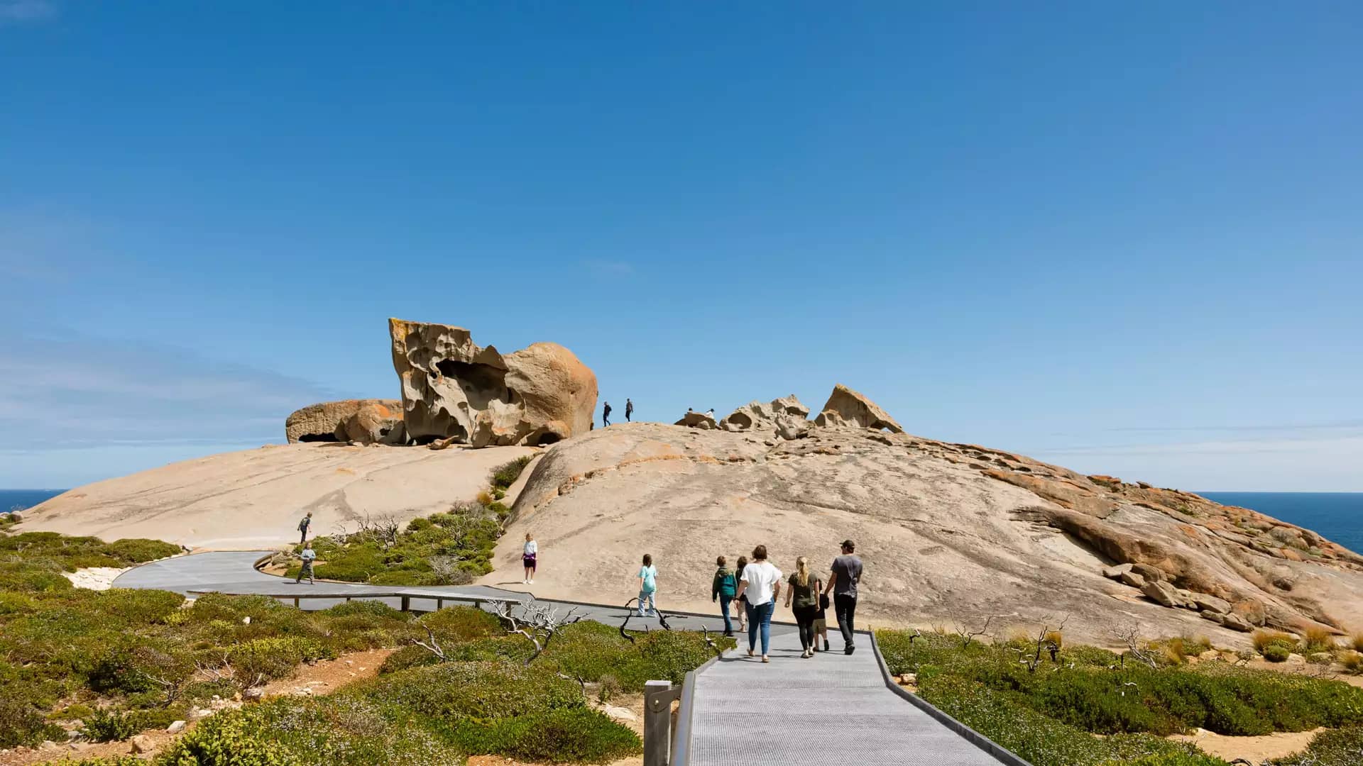Tourists walk along a wooden boardwalk towards huge orange lichen-covered granite boulders.