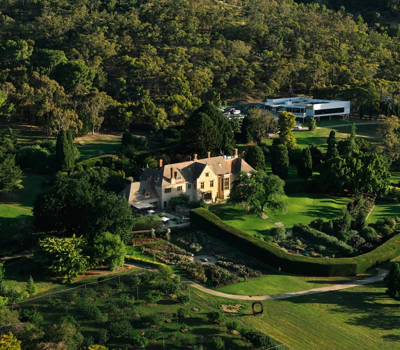An aerial view of a large sandstone manor house surrounded by lush formal gardens and dense trees.