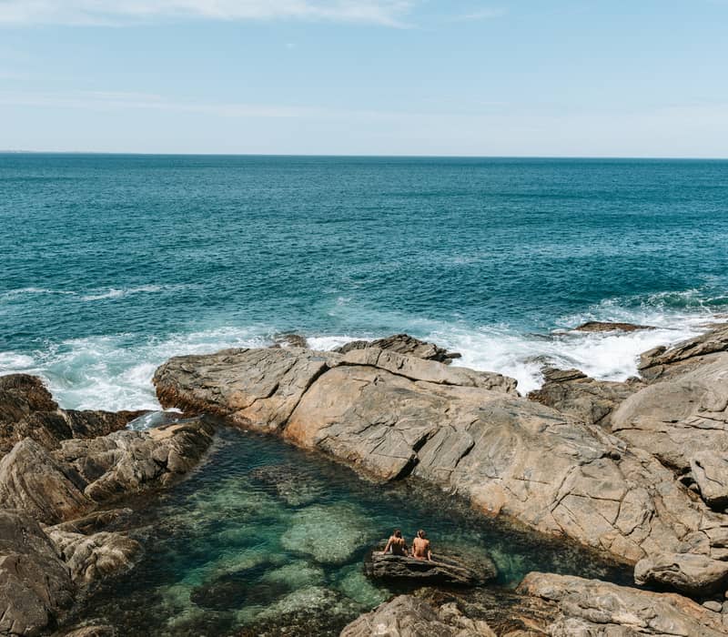 People sit in a clear, calm rock pool surrounded by jagged rocks with the ocean behind them.