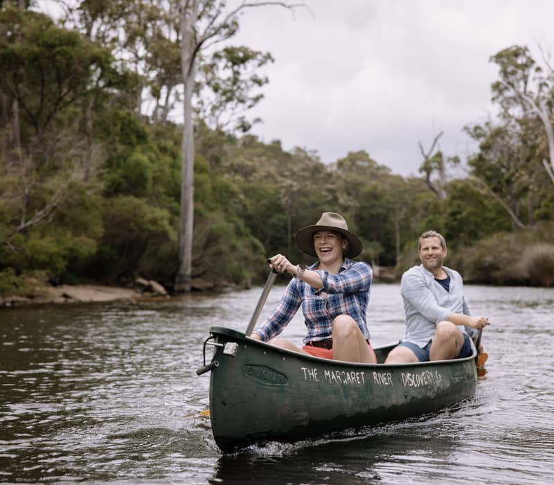 Two people in a green canoe paddling down a calm river surrounded by lush Australian bushland.