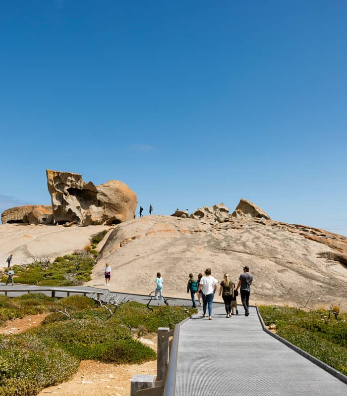 Tourists walk along a wooden boardwalk towards huge orange lichen-covered granite boulders.