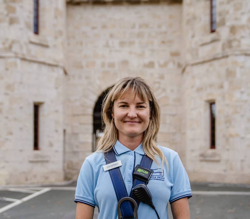 A female tour guide smiling in front of the large limestone gatehouse of the historic Fremantle Prison.