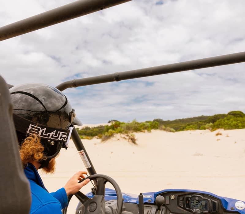 A person in a blue jacket and helmet drives a buggy through white sand dunes.