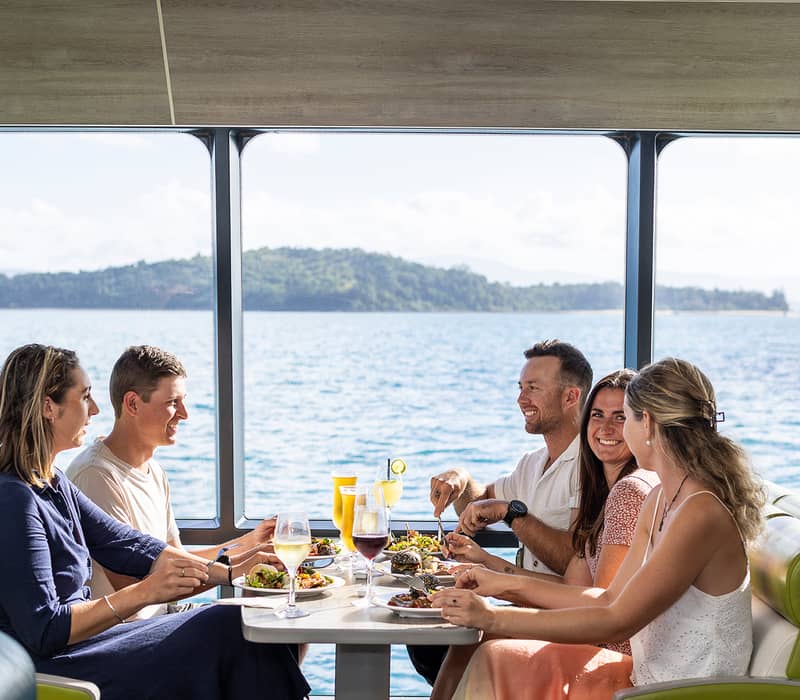 A group of friends dining on a boat with large windows overlooking the turquoise ocean in the Whitsundays.