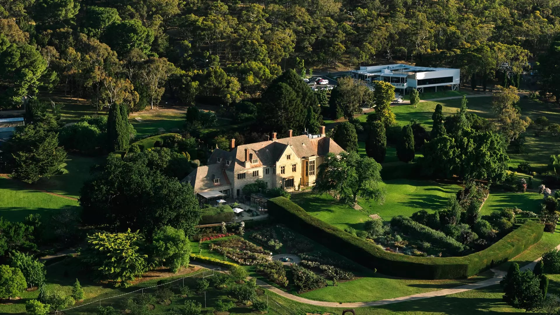 An aerial view of a large sandstone manor house surrounded by lush formal gardens and dense trees.