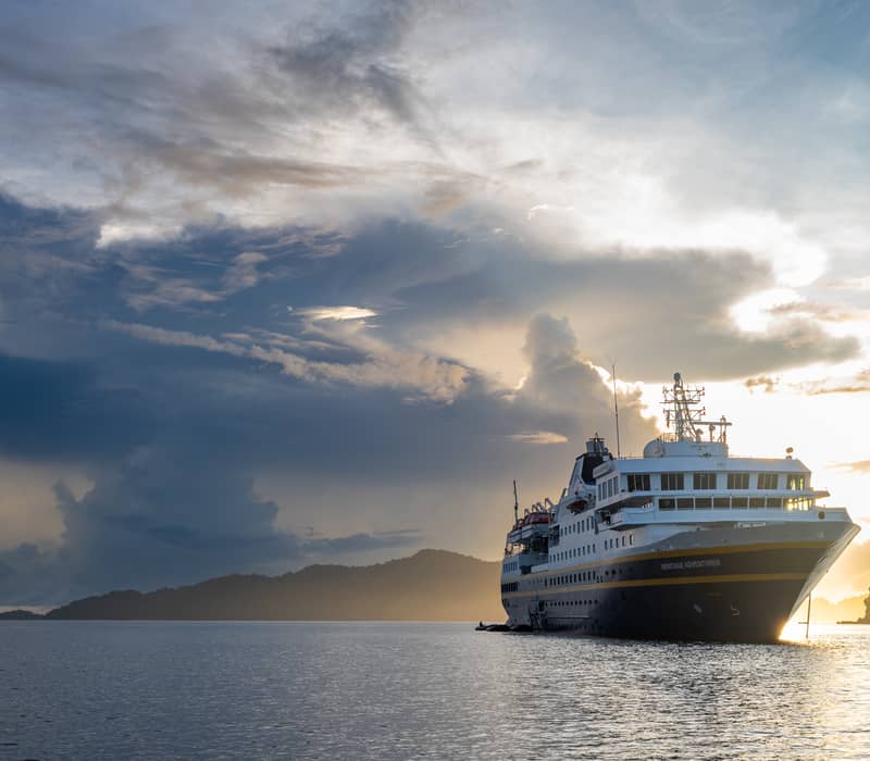 A sea-level shot of the Heritage Adventurer anchored in calm water. The sun is setting directly behind the ship, creating a brilliant golden light through dark, heavy clouds. A low, forested island is visible on the left.
