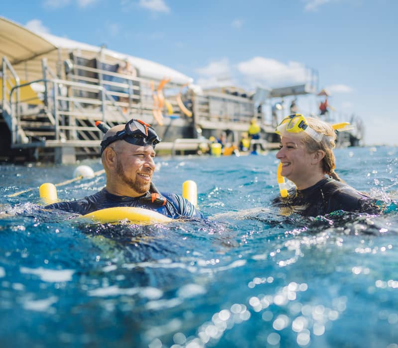 A man and woman wearing snorkeling gear smiling at each other while floating in the ocean next to a reef platform.