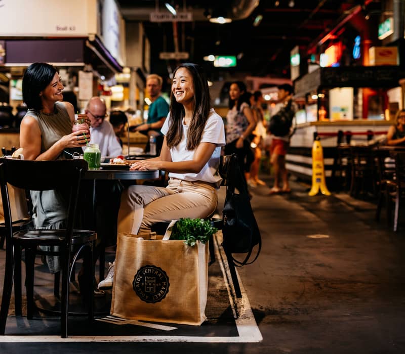 Two women laughing at a table with fresh smoothies and market bags in a vibrant indoor food market.