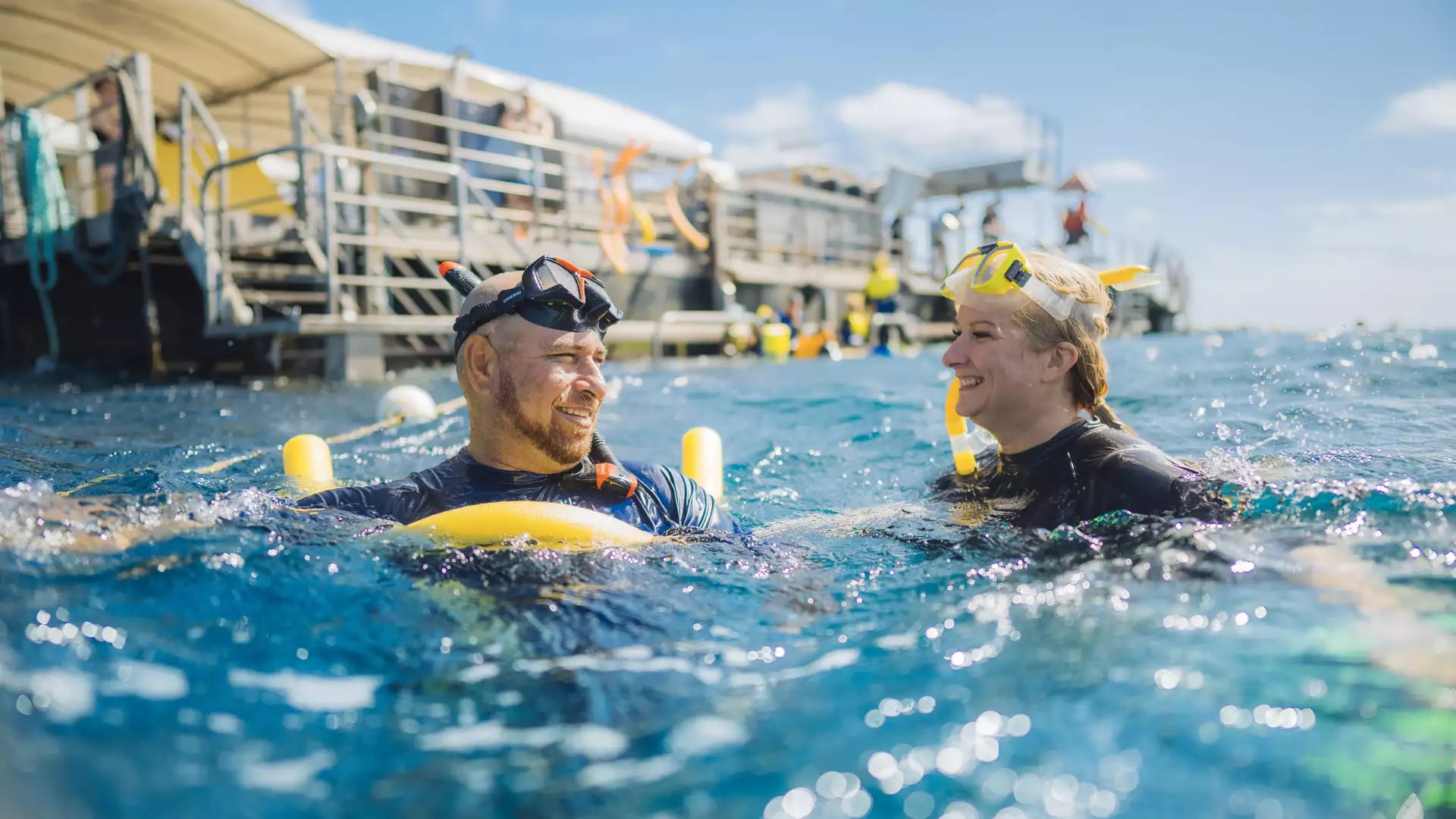 A man and woman wearing snorkeling gear smiling at each other while floating in the ocean next to a reef platform.