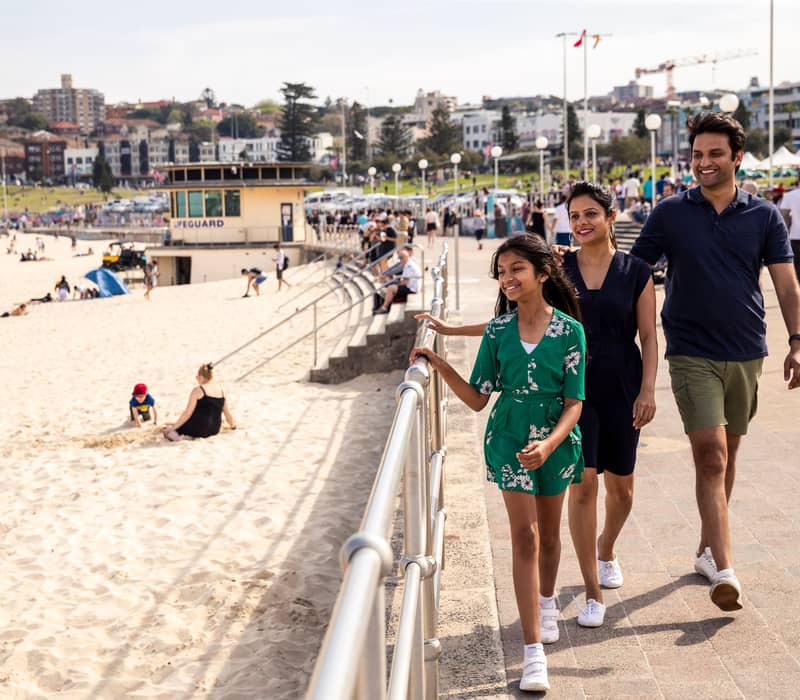 A family of three walking along the paved boardwalk at Bondi Beach on a sunny day in Sydney.