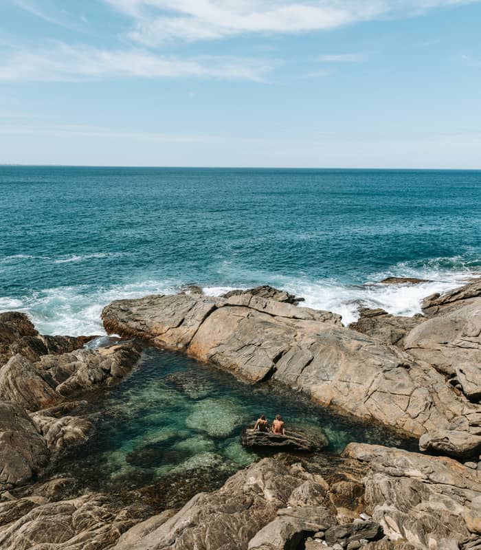 People sit in a clear, calm rock pool surrounded by jagged rocks with the ocean behind them.