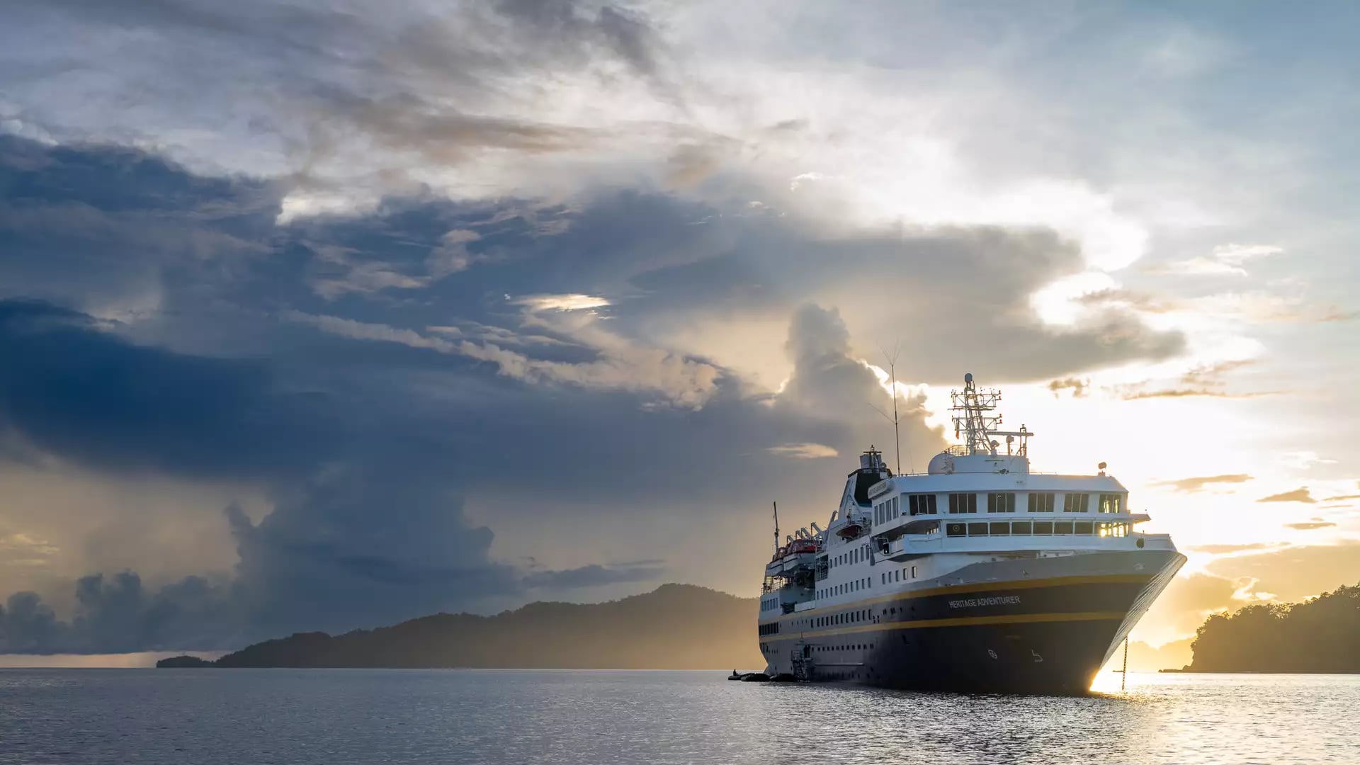A sea-level shot of the Heritage Adventurer anchored in calm water. The sun is setting directly behind the ship, creating a brilliant golden light through dark, heavy clouds. A low, forested island is visible on the left.