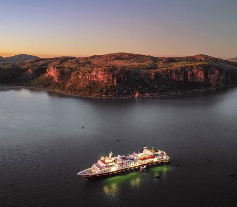 A high-angle aerial photograph at twilight showing the Heritage Adventurer cruise ship fully lit up, reflecting on calm dark water. Behind the ship is a large, dark orange-glowing cliff face under a soft purple and orange sunset sky.