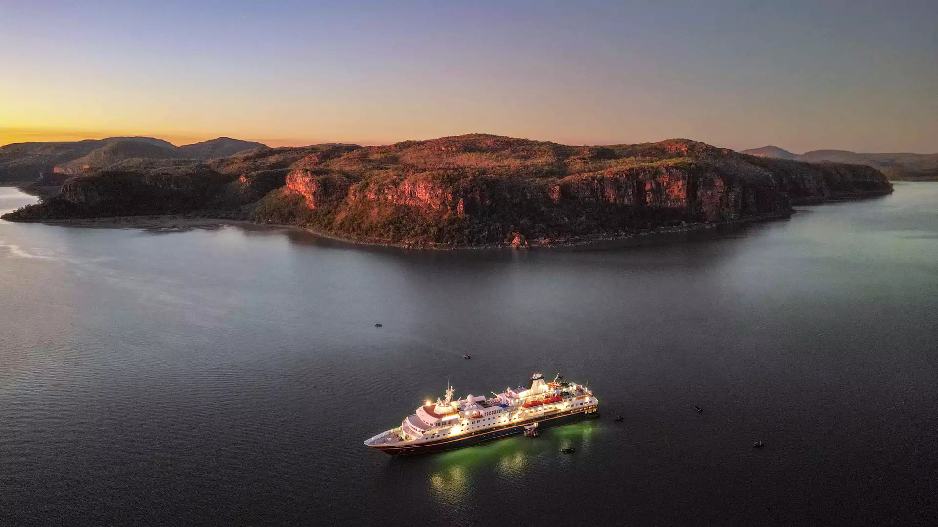 A high-angle aerial photograph at twilight showing the Heritage Adventurer cruise ship fully lit up, reflecting on calm dark water. Behind the ship is a large, dark orange-glowing cliff face under a soft purple and orange sunset sky.