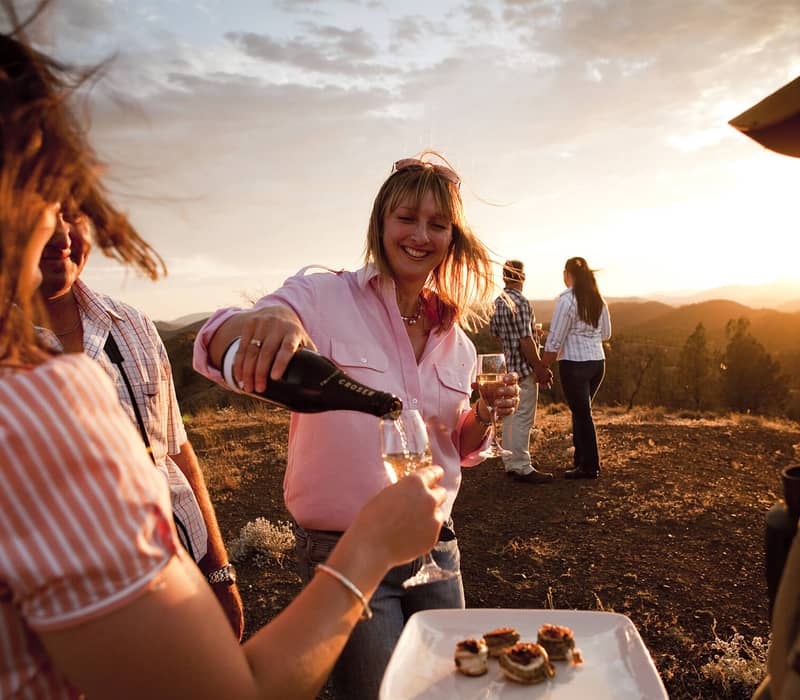A group of travelers enjoys sparkling wine and appetizers on a mountain ridge at sunset.