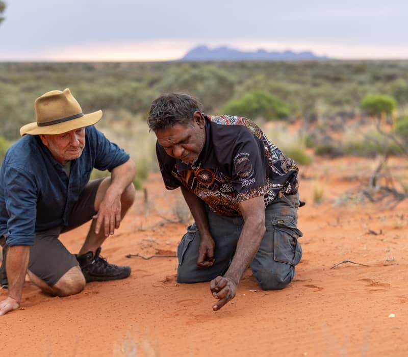 A guide and an Indigenous Australian man kneeling in the sand to look at tracks in the Northern Territory.
