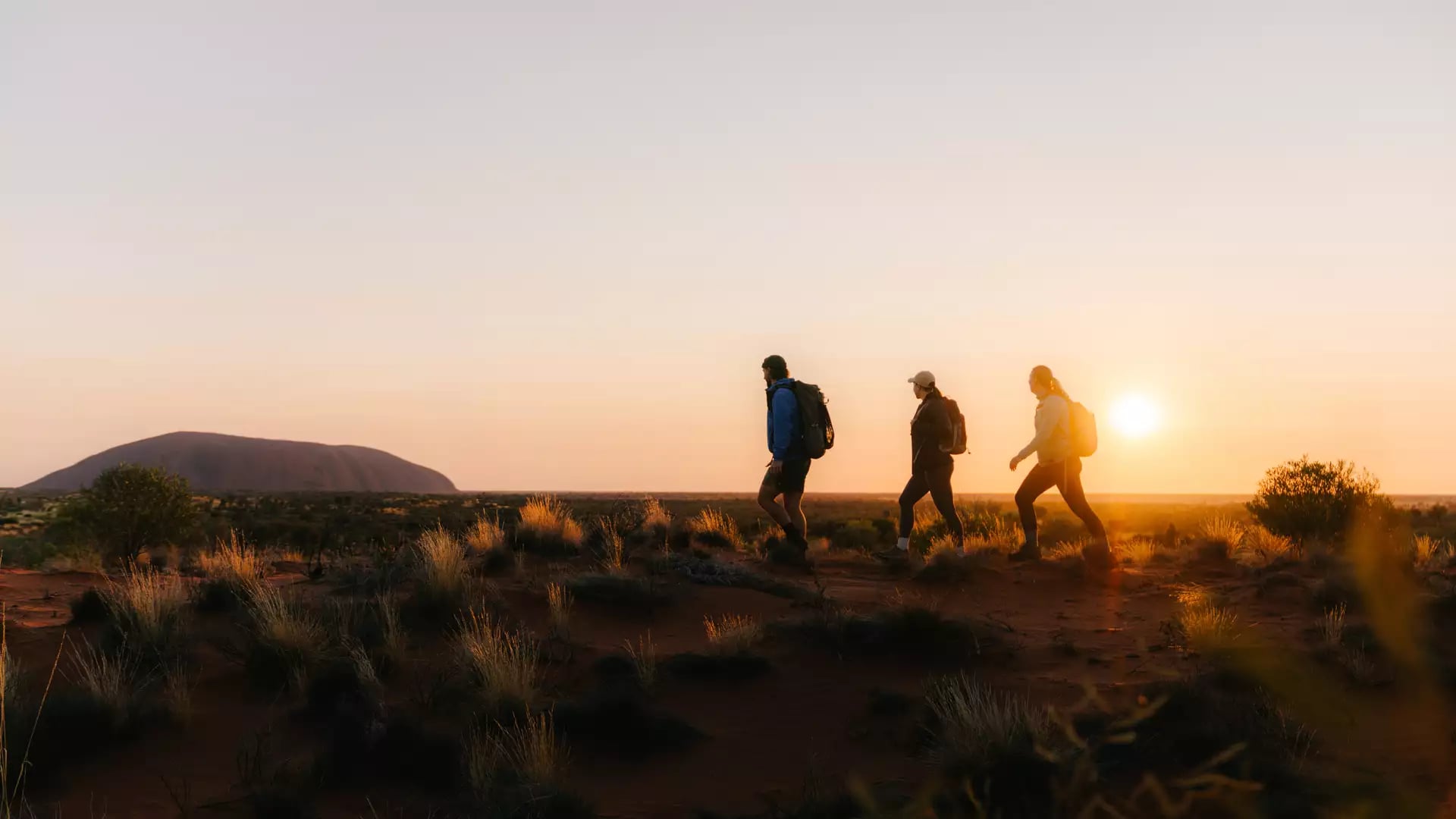 People hiking through the desert at sunrise with Uluṟu visible on the horizon.