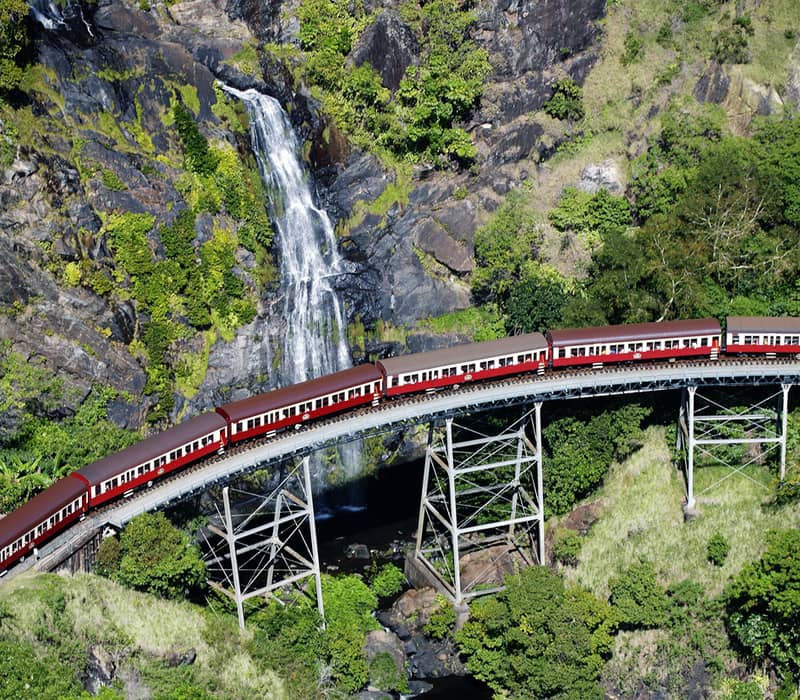 Vertical aerial view of a heritage train with red and beige carriages crossing a high trestle bridge beside a white waterfall in the forest.