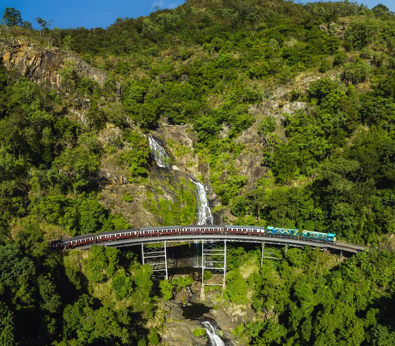 Aerial shot of a heritage train crossing a high trestle bridge in front of a multi-tiered waterfall in a lush green rainforest.