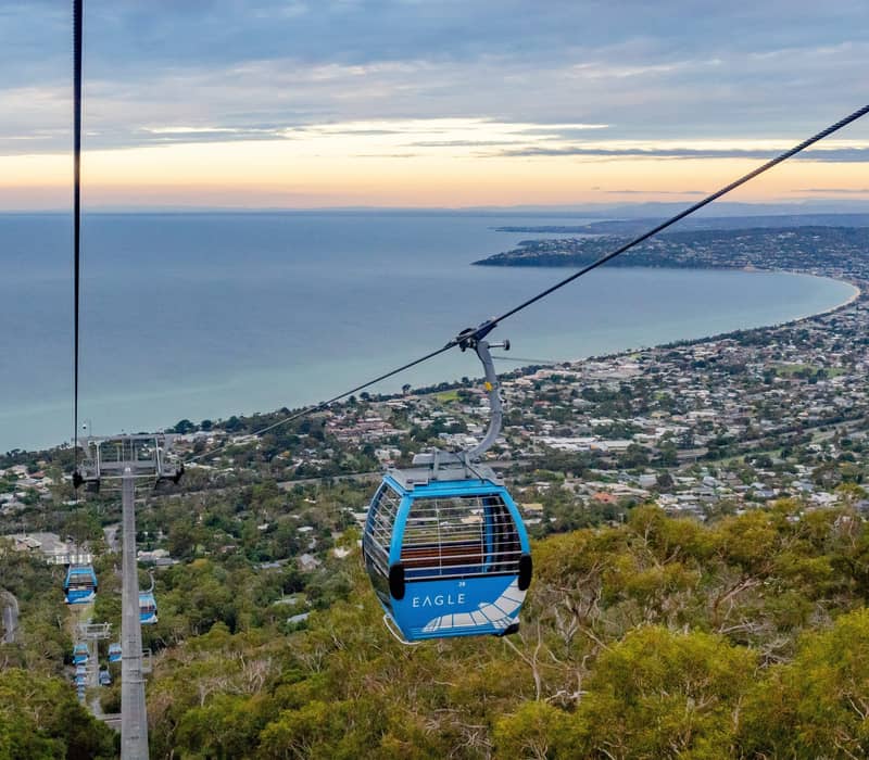 A blue gondola cabin hanging from a cable with a panoramic view of a bay and town at sunset.