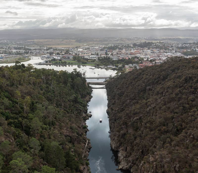 A high-angle aerial view of a narrow river gorge surrounded by dense forest leading to a city.