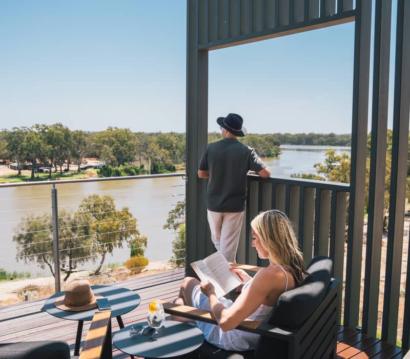 A woman sitting in a modern outdoor armchair reading a book while a man stands at the balcony railing looking out at a wide river landscape.