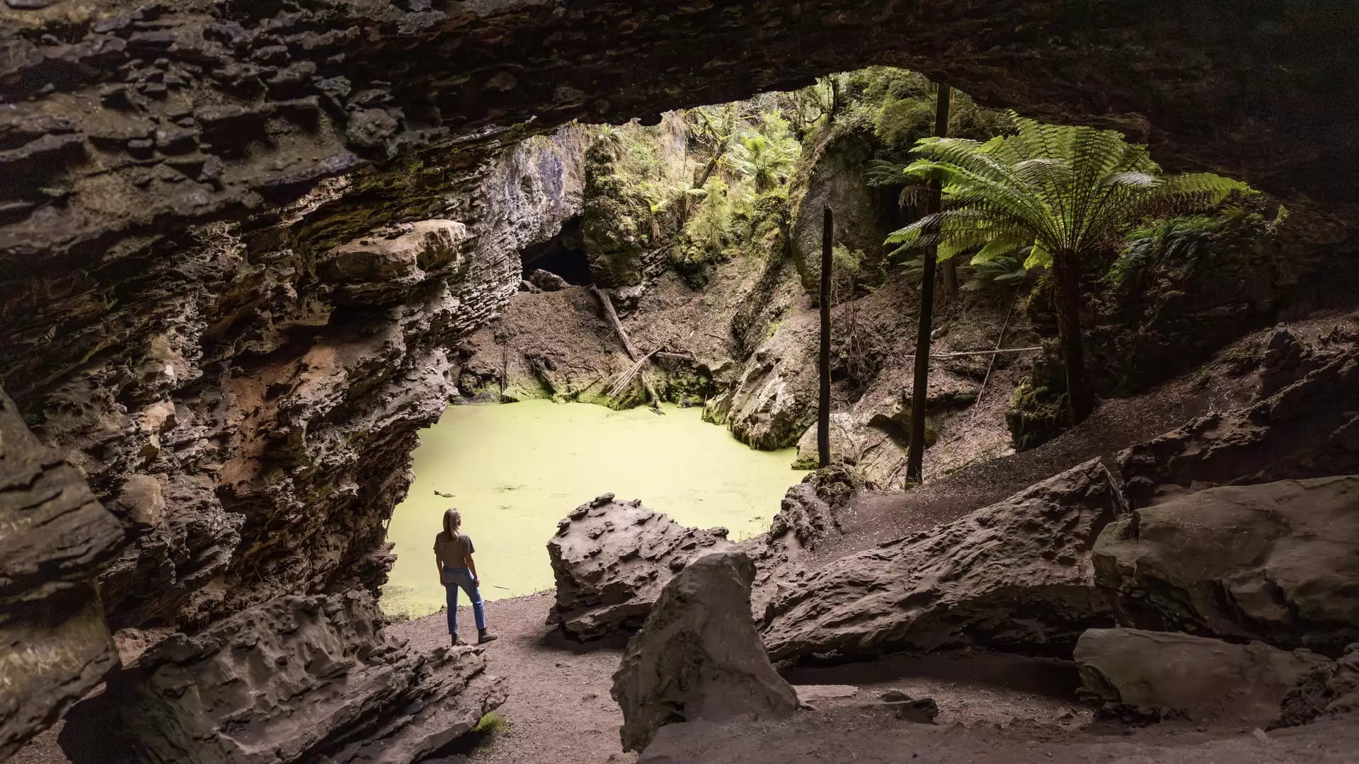 A person looking at a natural rock arch over a lime-green pond surrounded by rainforest.