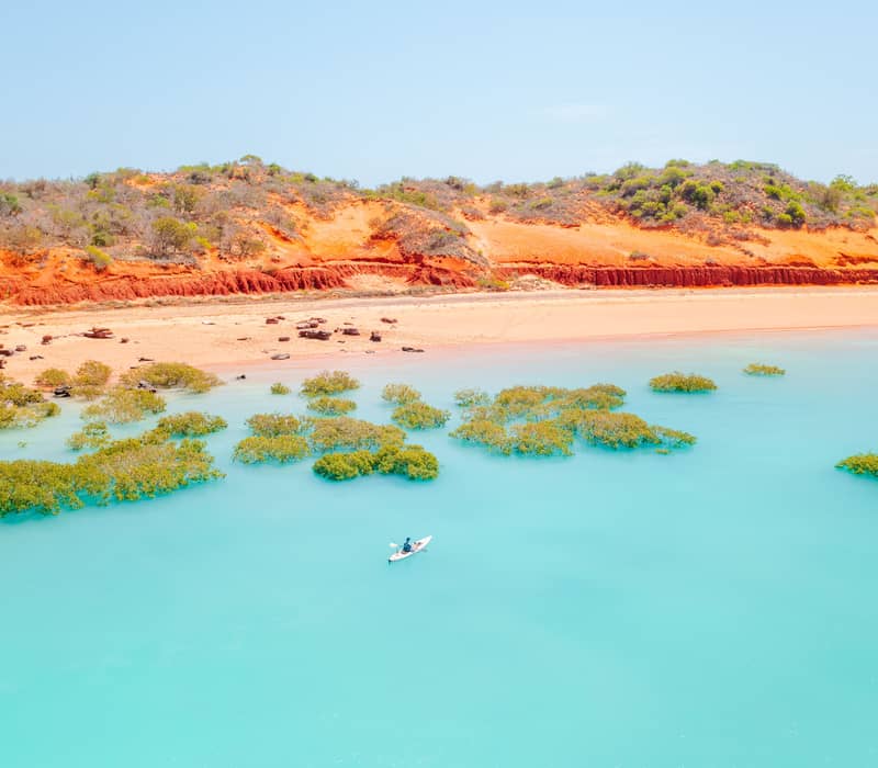 A lone kayaker paddles through the brilliant turquoise waters of Roebuck Bay, contrasted by vibrant red cliffs and green mangrove clusters.