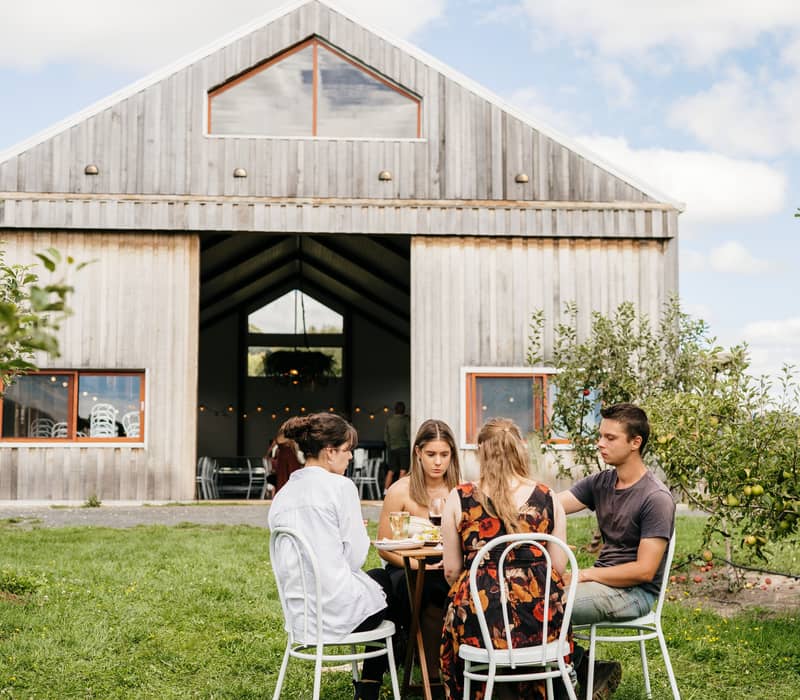 Four people sitting at an outdoor table eating and drinking in front of a large timber barn.