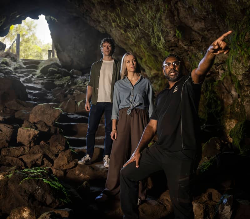 An Indigenous guide pointing upward while leading a tour through a rocky, moss-covered cave.