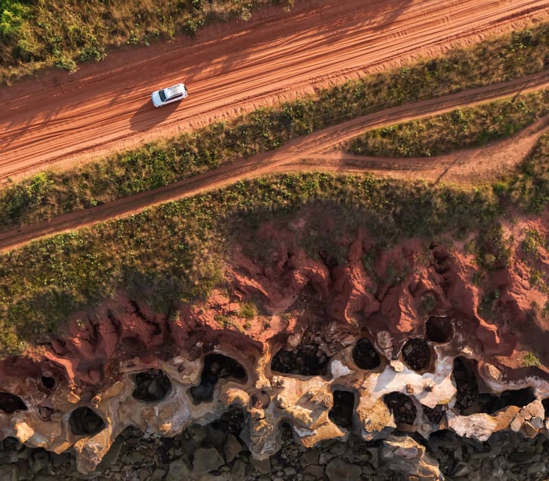 A striking top-down aerial view of a white 4WD vehicle navigating a red pindan track alongside the dramatic, eroded limestone cliffs of the Kimberley coastline.