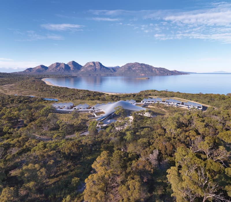 A breathtaking bird's-eye view of Saffire Freycinet, showcasing its stingray-inspired architectural roof nestled in the coastal bushland overlooking the Hazard Mountains and Wineglass Bay region.