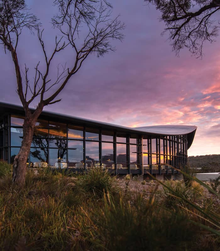 An external evening view of the Palate Restaurant at Saffire Freycinet, showcasing the iconic curved roof and floor-to-ceiling glass windows illuminated against a vibrant Tasmanian sunset.