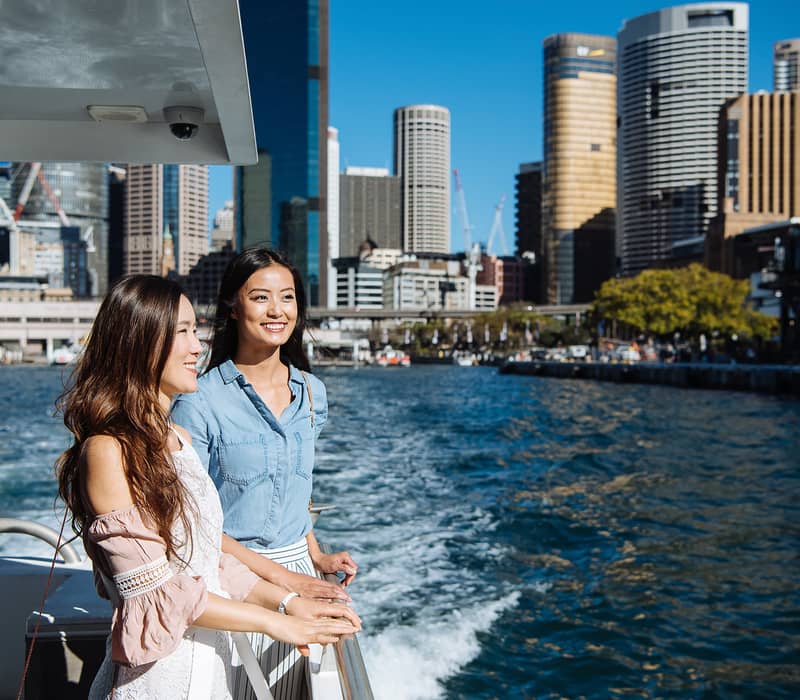 Two smiling women standing on the deck of a boat with the Sydney city skyscrapers and blue water behind them.