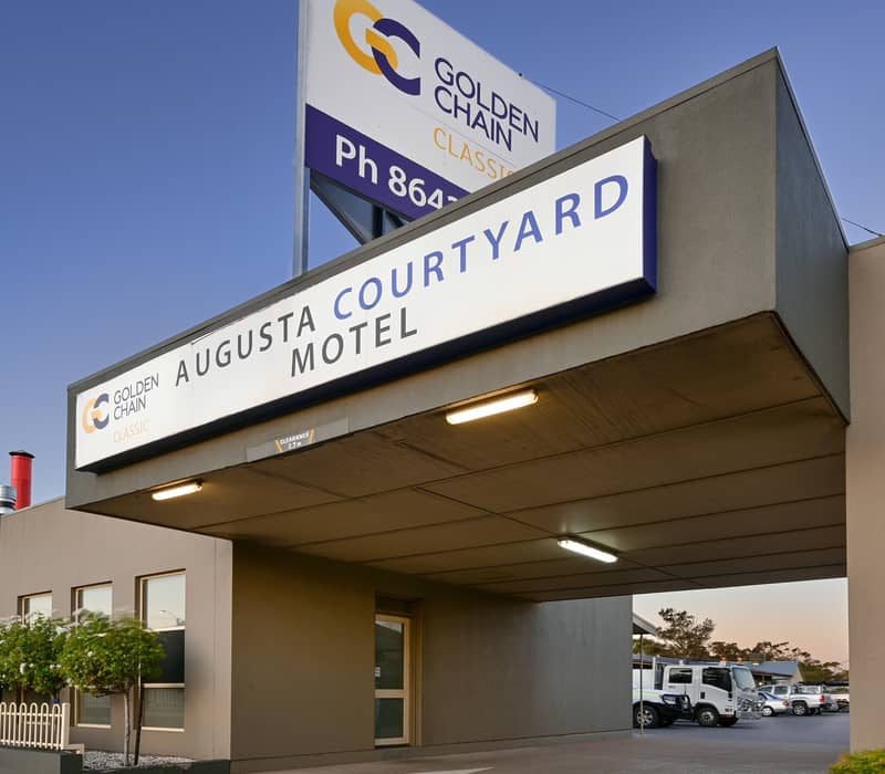Street-level view of a single-story motel entrance with a concrete overpass awning holding a white sign that reads "AUGUSTA COURTYARD MOTEL", topped by a tall rectangular "GOLDEN CHAIN" brand billboard against a deep blue sky.