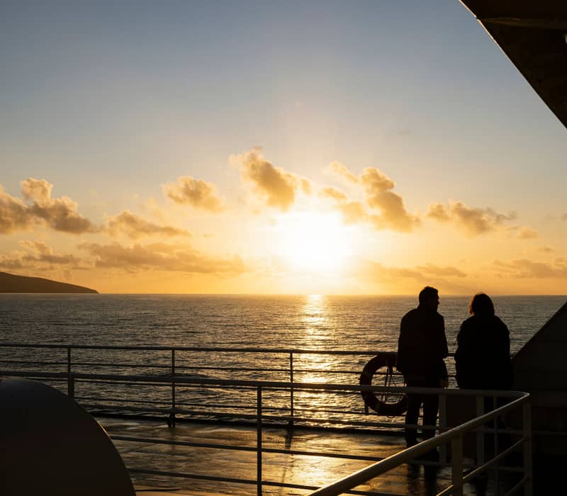 Silhouette of two people on a ferry deck looking out at a bright golden sunset over the ocean.