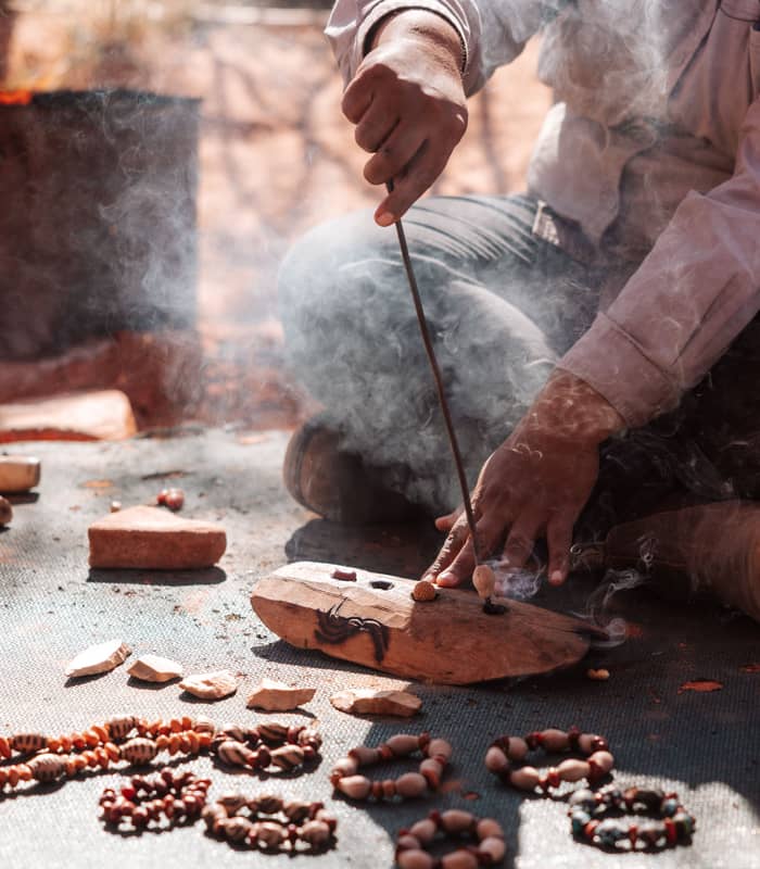 Close up of a person sitting on the ground using a metal tool to work on a piece of wood, with several finished seed bracelets displayed in the foreground.