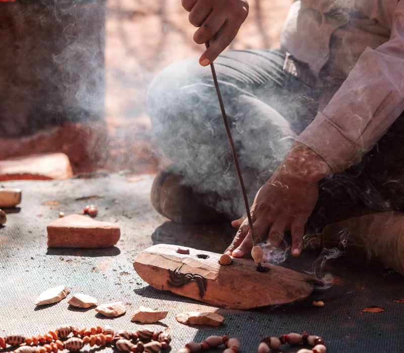 Close up of a person sitting on the ground using a metal tool to work on a piece of wood, with several finished seed bracelets displayed in the foreground.