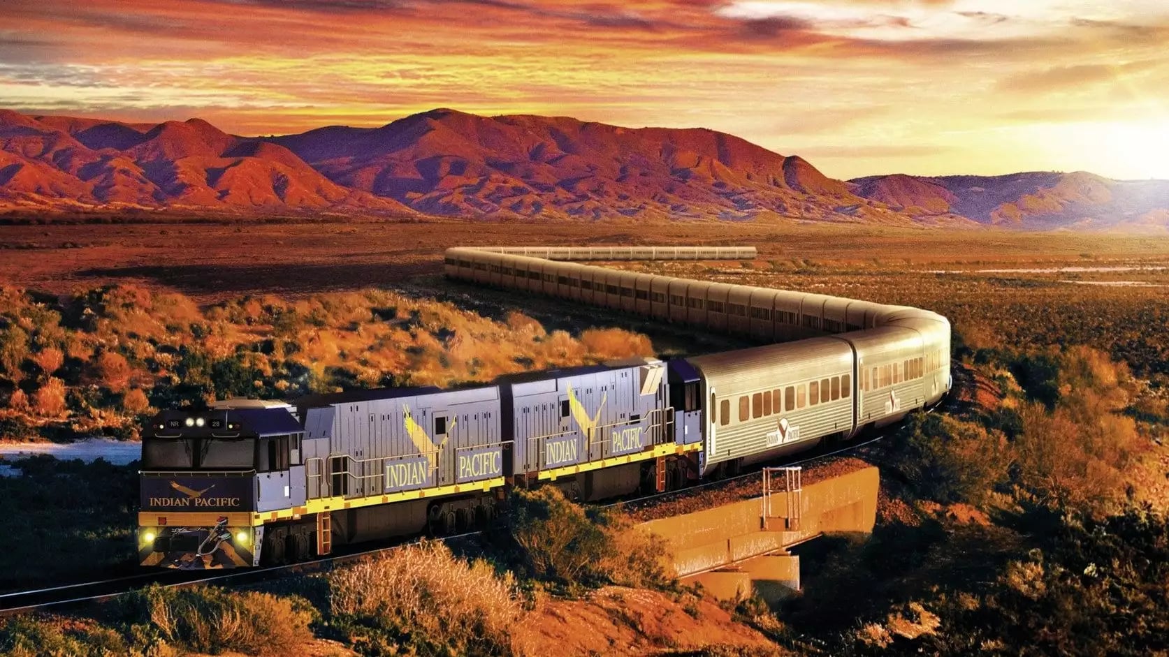 The Indian Pacific passenger train curves through the arid outback of South Australia under a dramatic orange and purple sunset sky.